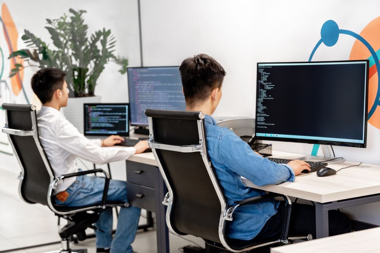 Offerings Two male developers at desks programming in a modern office workspace with large monitors.